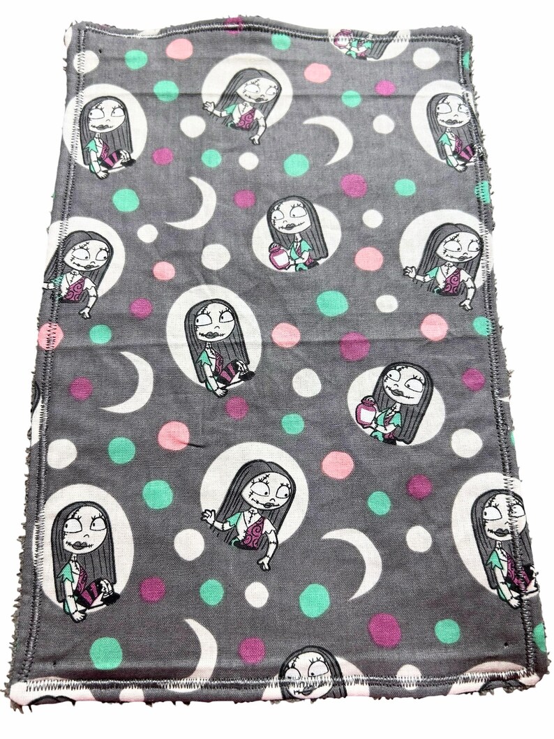 May include: Grey fabric featuring a repeating cartoon character design within white circles, interspersed with pink, teal, and white polka dots and crescent moons. The edges have a white stitched border.