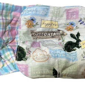 Può includere: Articolo in tessuto a tema pasquale con interno a quadri ed esterno stampato con un coniglietto, fiori e testo. L'esterno ha uno sfondo chiaro con venature del legno con colori pastello e le parole "Buona Pasqua", "Cotton Tail" e "Fresh Eggs".