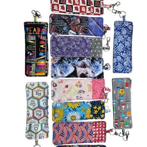 May include: A collection of eyeglass cases in various patterns and colors. Designs include a periodic table, floral prints, cat faces, and patriotic themes. Each case has a keychain clip and some feature the word "READ".