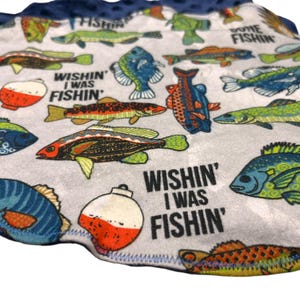 May include: A light gray, patterned fabric featuring colorful illustrations of various fish species and fishing bobbers. The phrase "Wishin' I Was Fishin'" is printed in black. The fabric has a dark blue, scalloped edge.