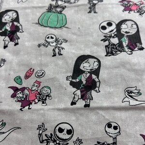 May include: A light grey fabric patterned with cartoon characters from The Nightmare Before Christmas. The design includes Jack Skellington, Sally, and other characters in black, white, and shades of purple, pink, and green.