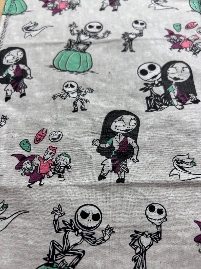 May include: A light grey fabric patterned with cartoon characters from The Nightmare Before Christmas. The design includes Jack Skellington, Sally, and other characters in black, white, and shades of purple, pink, and green.