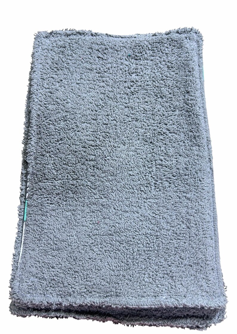 May include: A folded, grey, plush towel with a textured surface. The towel has a soft, absorbent appearance, suggesting it is made of a high-quality material. The edges are neatly finished, and a white and green accent is visible.