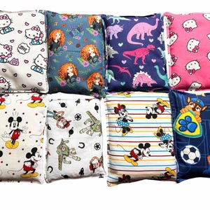 May include: A collection of eight rectangular fabric pillows featuring various character prints including Hello Kitty, Merida, dinosaurs, Mickey Mouse, Toy Story characters, Minnie Mouse, and Paw Patrol, displayed in a grid arrangement.