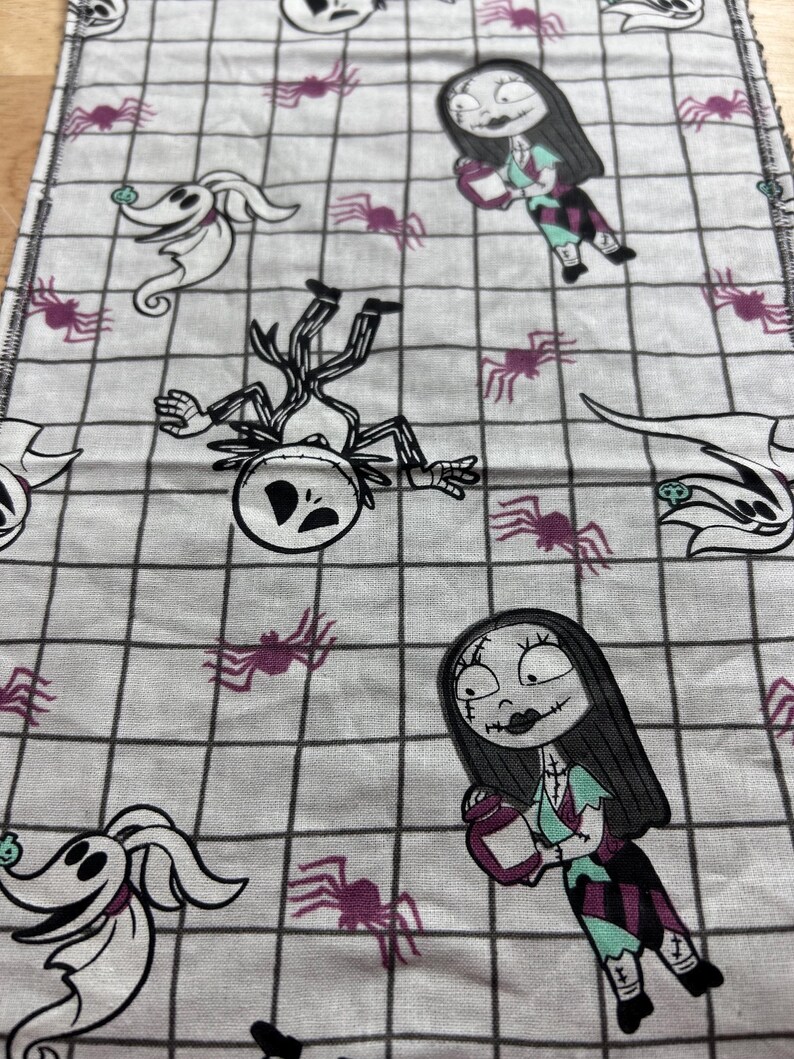 May include: White fabric with a black grid pattern, featuring characters from The Nightmare Before Christmas. The design includes Jack Skellington, Sally, Zero, and purple spiders. Suitable for crafting or decorative purposes.
