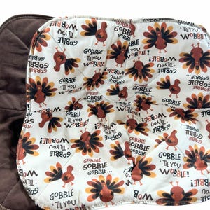 May include: A brown and white Thanksgiving-themed quilted pot holder. The white side features a repeating pattern of cartoon turkeys and the text "GOBBLE 'TIL YOU WOBBLE!". The reverse side is a solid brown fabric.