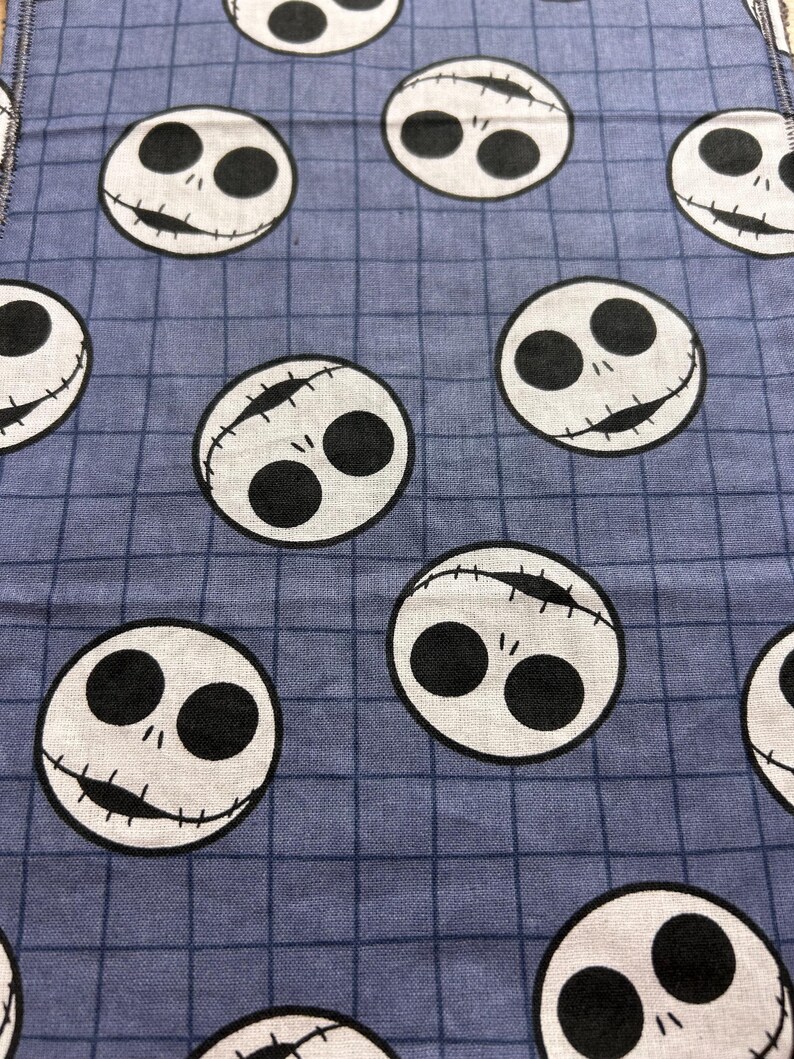 May include: Close-up of a blue fabric with a grid pattern. The fabric is covered with white circles featuring black eyes and stitched mouths, resembling a character from a popular animated film. The design is repeated across the fabric.