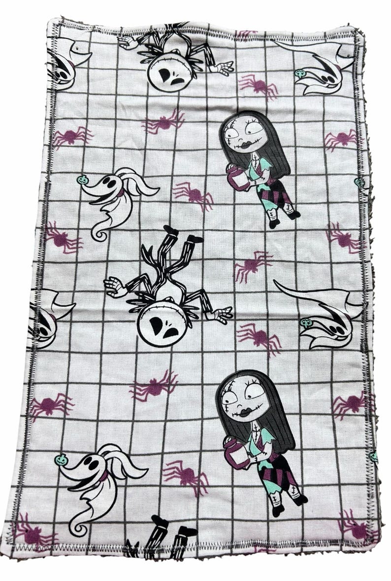 May include: White fabric with a black grid pattern featuring cartoon characters from The Nightmare Before Christmas. The design includes Jack Skellington, Sally, Zero, and purple spiders. The edges are finished with a black zigzag stitch.