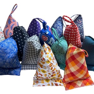 May include: A collection of various fabric doorstops in assorted patterns and colors. Designs include floral, polka dots, gingham, and patriotic themes. Each doorstop has a loop handle for easy carrying.