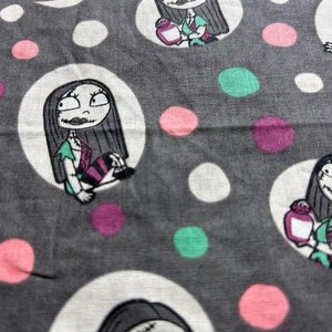 May include: Grey fabric featuring a repeating pattern of Sally from The Nightmare Before Christmas. Sally is in a white circle, set against a grey backdrop, with white, pink, teal, and purple polka dots and a white crescent moon.