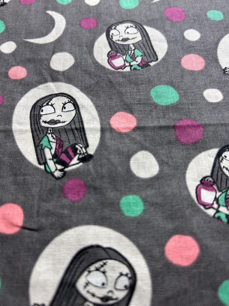 May include: Grey fabric featuring a repeating pattern of Sally from The Nightmare Before Christmas. Sally is in a white circle, set against a grey backdrop, with white, pink, teal, and purple polka dots and a white crescent moon.