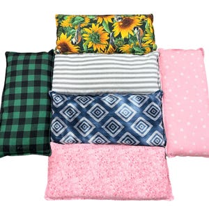 May include: A selection of rectangular pillows featuring diverse patterns. Designs include sunflowers, green plaid, blue diamond shapes, pink floral, grey and white stripes, and pink with white dots. The pillows are arranged in a stacked, fan-like display.