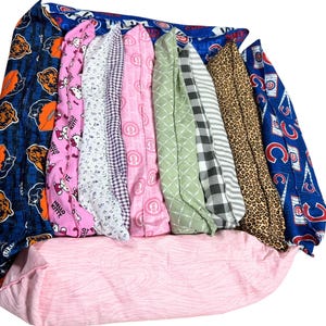 Könnte beinhalten: Eine Sammlung farbenfroher Stoffschläuche, jeder mit einem einzigartigen Muster. Zu den Designs gehören Fußbälle, Hello Kitty, Blumenmuster, Vichy-Karo und Tiermuster. Die Stoffe sind auf einer rosa gemusterten Oberfläche angeordnet, mit einem blau gemusterten Stoff oben.