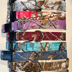 Op de afbeelding: Een collectie van negen hondenhalsbanden met diverse Realtree camouflagepatronen in kleuren zoals zwart, wit, roze, paars, rood, lichtblauw, donkerblauw, beige en oranje, elk voorzien van een zwarte kunststof gesp en een metalen D-ring.