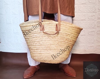 May include: A large, woven straw tote bag with brown leather handles. The bag is being held by a person wearing a brown tunic and white pants.