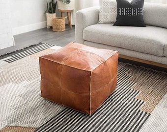 May include: A brown leather pouf ottoman with a square shape and stitched panels sits on a black and white striped rug in a living room setting.