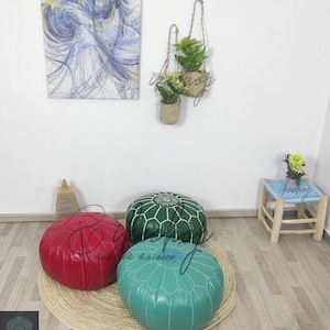 May include: Three poufs, one red, one green, and one turquoise blue, are arranged on a woven straw mat. The poufs are all round and have a tufted design. The poufs are in a room with a wooden floor and a white wall. There are two hanging plants in woven baskets on the wall.