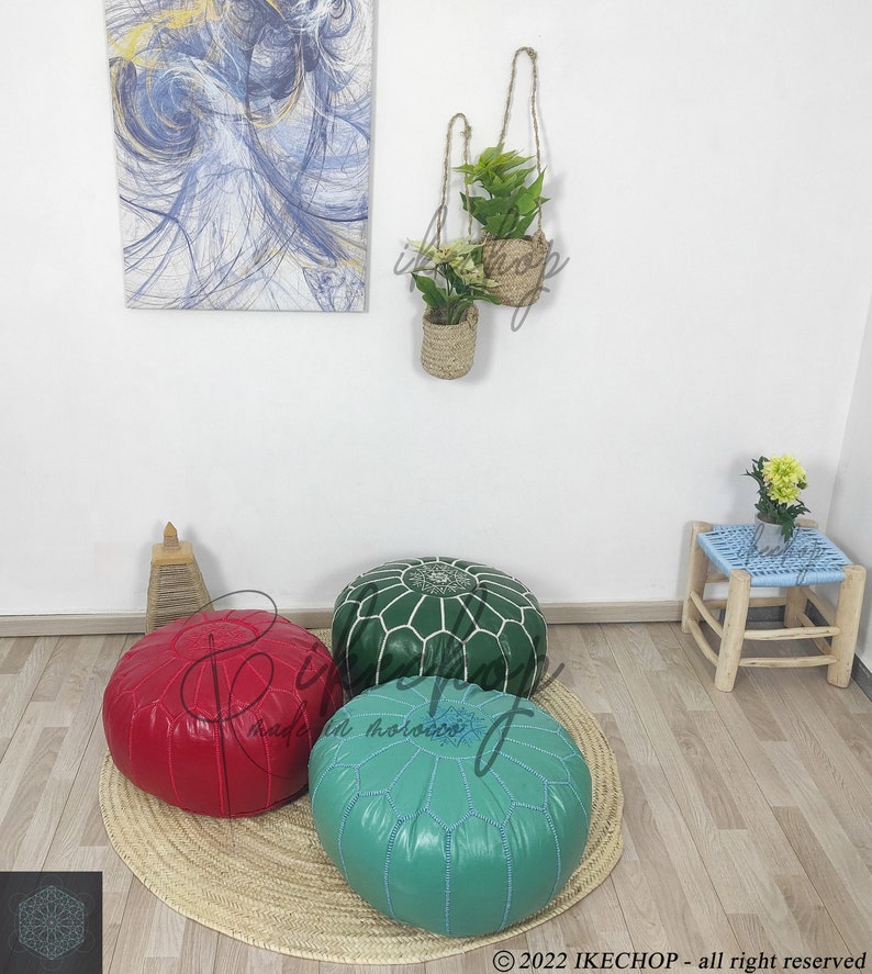 May include: Three poufs, one red, one green, and one turquoise blue, are arranged on a woven straw mat. The poufs are all round and have a tufted design. The poufs are in a room with a wooden floor and a white wall. There are two hanging plants in woven baskets on the wall.