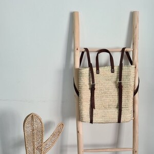 May include: A natural woven straw bag with brown leather straps. The bag is hanging on a wooden ladder.