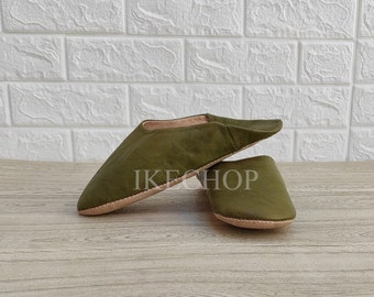 May include: Olive green leather slippers with a pointed toe and a rounded heel. The slippers are displayed on a wooden surface with the text "IKE SHOP" visible. The slippers have a light brown sole.