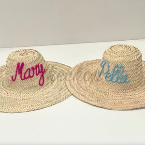 May include: Two straw sun hats with the names "Mary" and "Bella" embroidered on them in pink and blue thread.