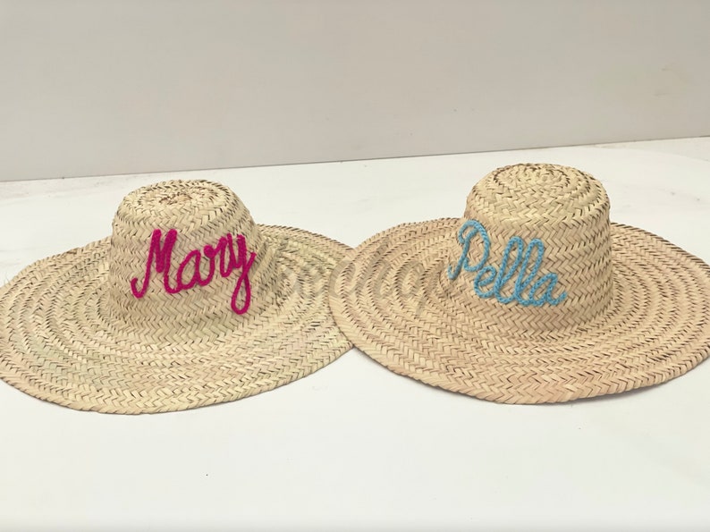 May include: Two straw sun hats with wide brims. One hat has the name "Mary" embroidered in pink, and the other has "Pella" in blue. The hats are made of woven straw and are suitable for sun protection.