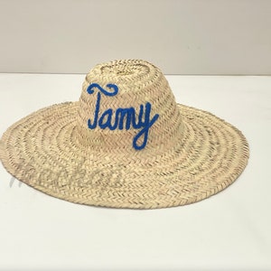 May include: A woven straw sun hat with a wide brim. The word "Tamy" is embroidered in blue cursive across the crown. The hat is a natural beige color and appears to be handmade.