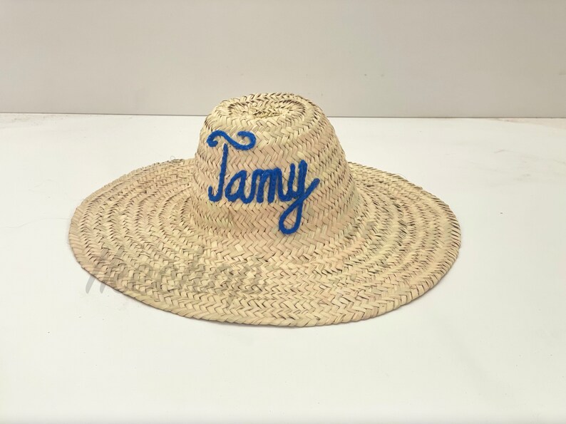 May include: A woven straw sun hat with a wide brim. The word "Tamy" is embroidered in blue cursive across the crown. The hat is a natural beige color and appears to be handmade.