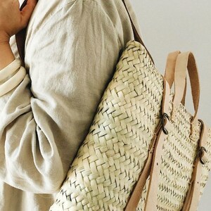 May include: A woven straw backpack with brown leather straps. The backpack is being worn by a person wearing a light beige shirt.