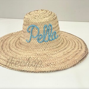 May include: A wide-brimmed straw sun hat with the name "Pella" embroidered in light blue thread across the crown. The hat features a woven design and a wide brim for sun protection. A stylish accessory for sunny days.