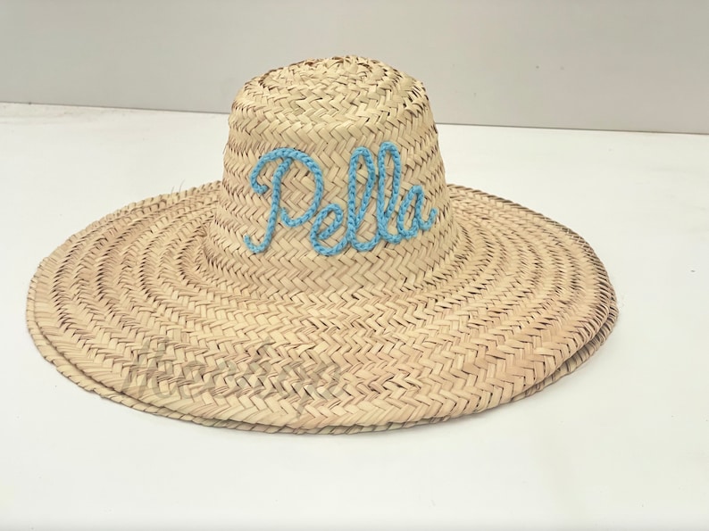 May include: A wide-brimmed straw sun hat with the name "Pella" embroidered in light blue thread across the crown. The hat features a woven design and a wide brim for sun protection. A stylish accessory for sunny days.