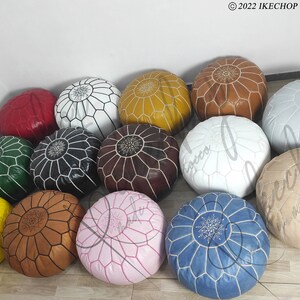 May include: A collection of 14 colorful poufs, each with a unique geometric pattern and a white embroidered design in the center. The poufs are made of leather and are arranged in a circle on a wooden floor.