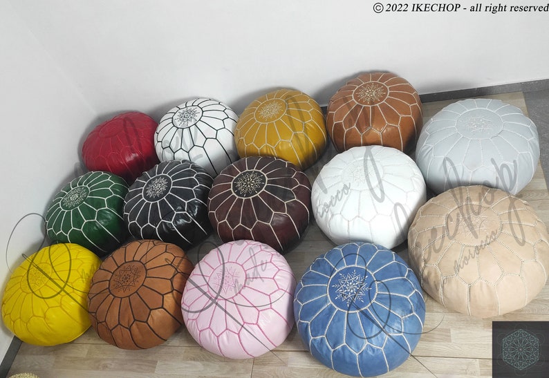 May include: A collection of 14 colorful poufs, each with a unique geometric pattern and a white embroidered design in the center. The poufs are made of leather and are arranged in a circle on a wooden floor.