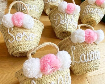 Könnte beinhalten: Eine Sammlung kleiner Strohkörbe mit weißen und rosa Pompons. Jeder Korb hat einen Namen in weißer Stickerei. Die Namen sind: 'Sofia', 'Delphine', 'Laura', 'Louise', 'Marine', 'Mama' und 'Malla'.