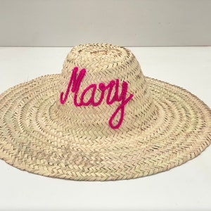 May include: A woven straw sun hat with a wide brim. The word "Mary" is embroidered across the crown in bright pink yarn. The hat is made of natural-colored straw.