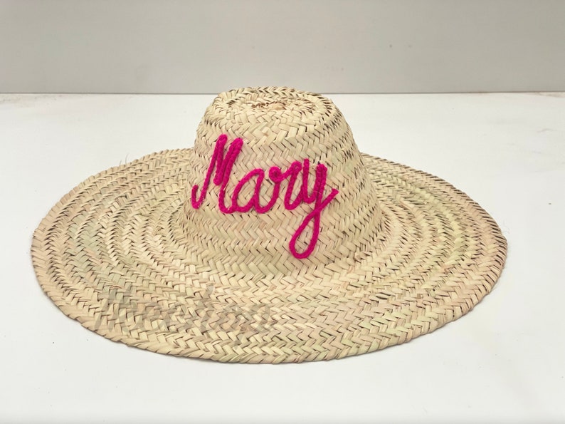 May include: A woven straw sun hat with a wide brim. The word "Mary" is embroidered across the crown in bright pink yarn. The hat is made of natural-colored straw.