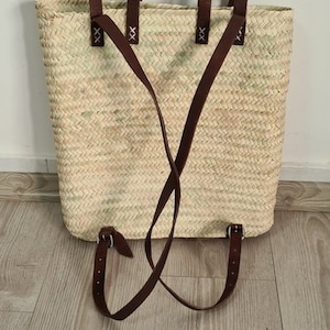 May include: A woven straw tote bag with brown leather straps and buckles. The bag has a top handle and adjustable straps that can be worn as a backpack.