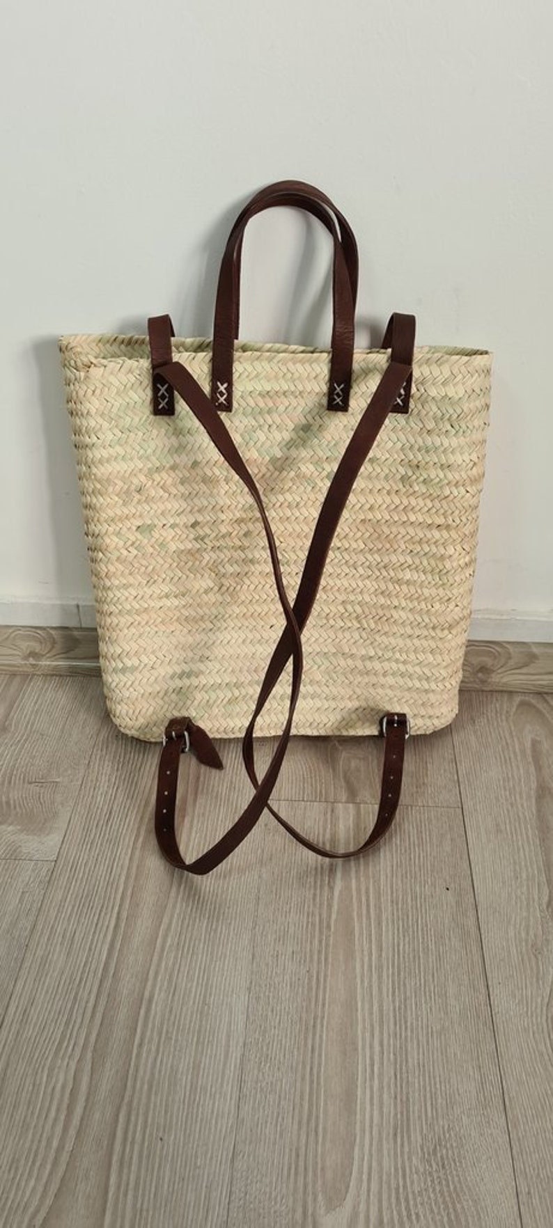 May include: A woven straw tote bag with brown leather straps and buckles. The bag has a top handle and adjustable straps that can be worn as a backpack.