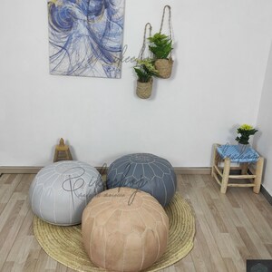 May include: Three poufs in different colors: gray, beige, and blue. The poufs are made of leather and have a tufted design. They are sitting on a woven rug in a room with a wooden floor.