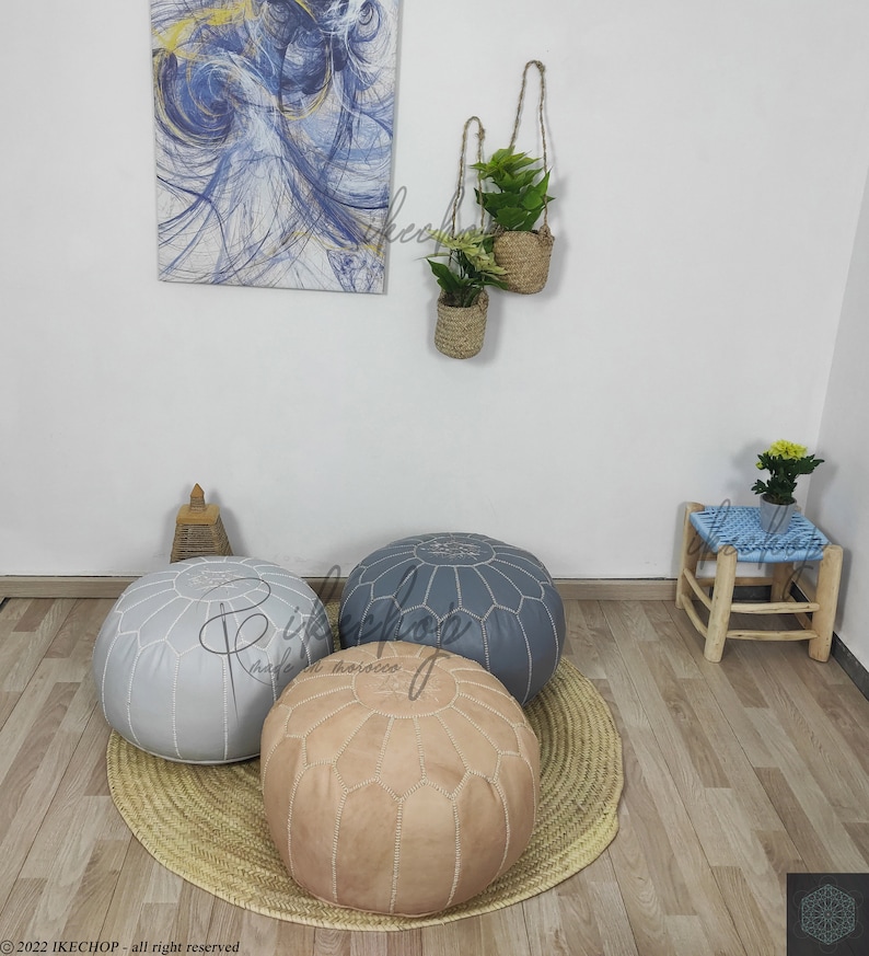 May include: Three poufs in different colors: gray, beige, and blue. The poufs are made of leather and have a tufted design. They are sitting on a woven rug in a room with a wooden floor.
