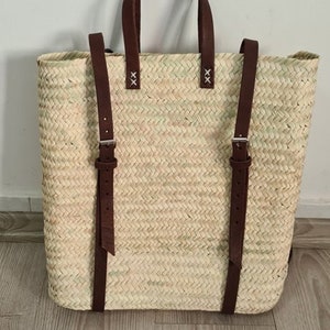 May include: A woven straw tote bag with brown leather straps and buckles. The bag has a top handle and two adjustable shoulder straps.