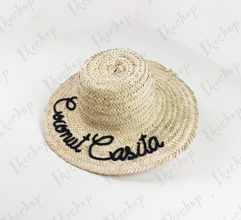 May include: A natural straw hat with a wide brim and a rounded crown. The hat features the embroidered text "Coconut Casita" in black cursive lettering across the front. The hat is designed for sun protection.