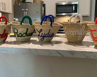 Könnte beinhalten: Fünf Strohtaschen mit bunten Henkeln und personalisierten Namen in gestickten Buchstaben. Die Namen sind "Lydie", "Jennifer", "Rachael", "T" und "Andy".