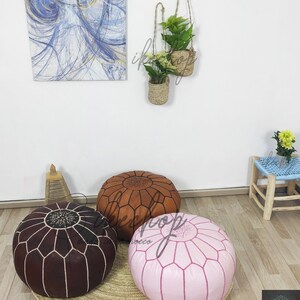 May include: Three round leather poufs in brown, pink, and tan with intricate stitching. The poufs are on a woven rug.