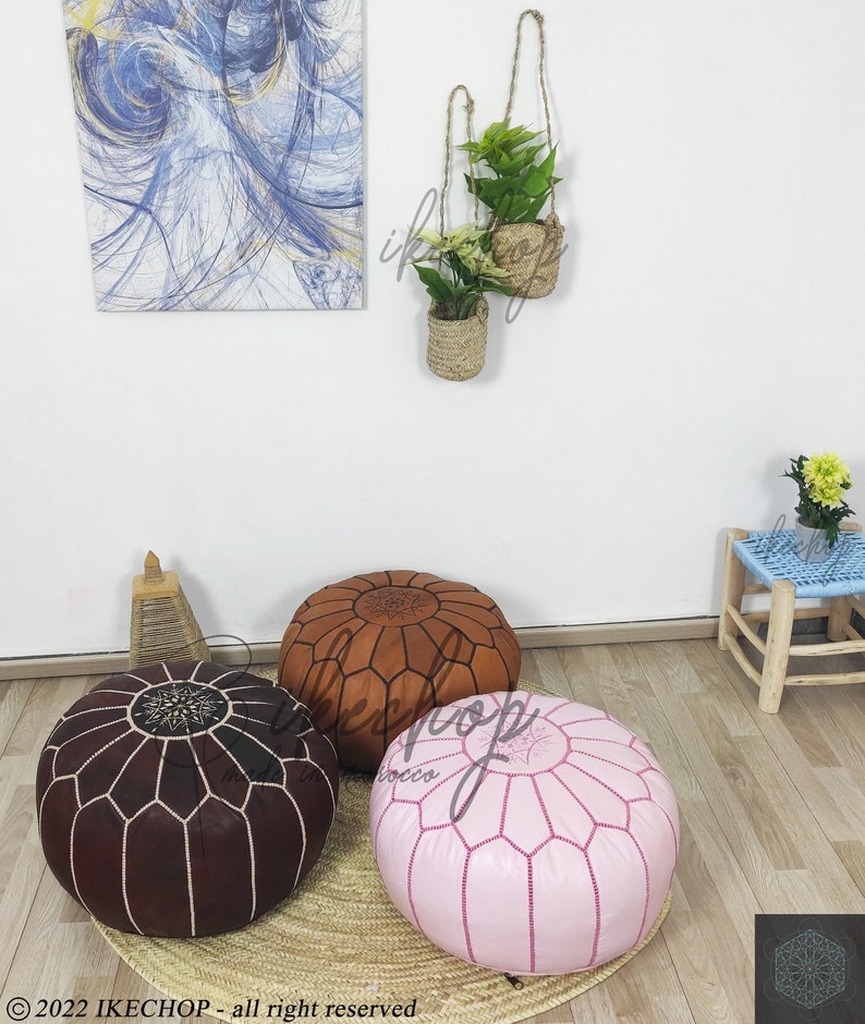 May include: Three round leather poufs in brown, pink, and tan with intricate stitching. The poufs are on a woven rug.