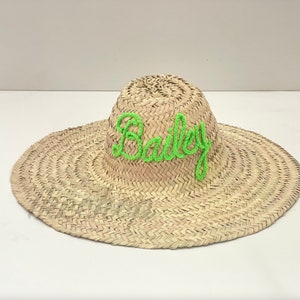 May include: A wide-brimmed straw hat with the name "Bailey" embroidered in bright green thread across the crown. The hat is made of woven straw and has a natural, light brown color.