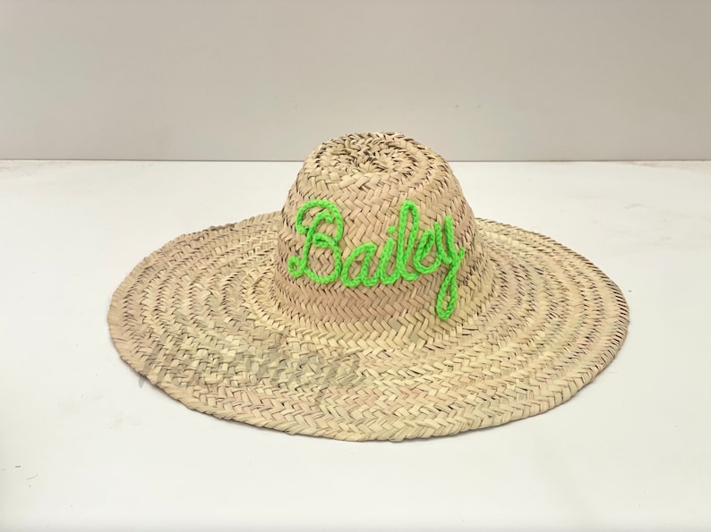 May include: A wide-brimmed straw hat with the name "Bailey" embroidered in bright green thread across the crown. The hat is made of woven straw and has a natural, light brown color.