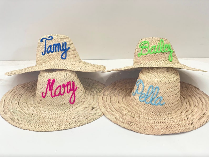 May include: Four straw sun hats with wide brims. Each hat has a different name embroidered on the crown in bright colors: Tamy (blue), Mary (pink), Bailey (green), and Della (light blue). The hats are displayed on a white surface.