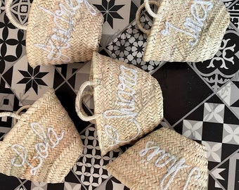 May include: Five straw tote bags with white embroidered lettering. The bags are on a black and white tiled surface. The lettering on the bags reads "Lola", "Sofia", "Jules", "Alexa", and "Scout".