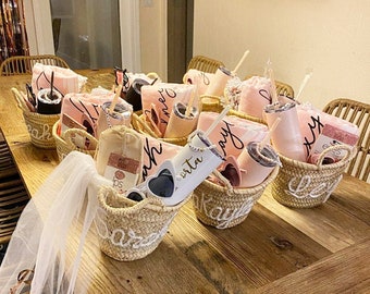 May include: A collection of small wicker baskets filled with gifts for a bridal shower. Each basket contains a pink towel, a tumbler with a straw, sunglasses, and a veil. The baskets are labeled with the names of the bridesmaids.